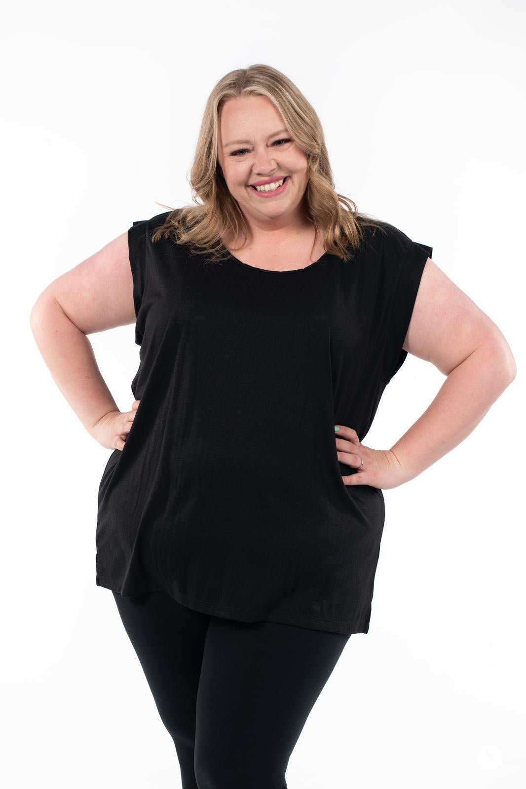 Smiling plus-size woman modeled in black Cap Sleeve Scoop Neck Tee by SweetTops with hands on hips on white backdrop