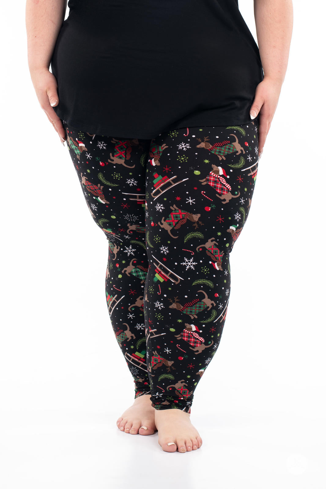 Plus-size model wearing SweetLegs Happy Pawlidays leggings with Dachshunds in holiday plaid and festive sled designs on black background