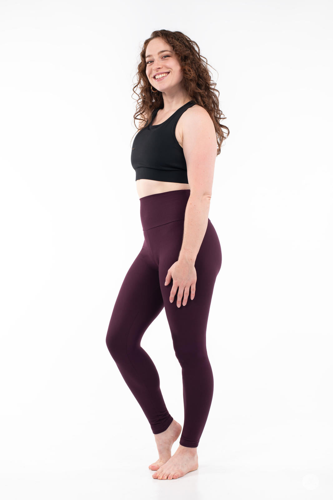 Smiling model in SweetLegs Blackberry High-Waisted Leggings in deep rich purple with black crop top side view barefoot