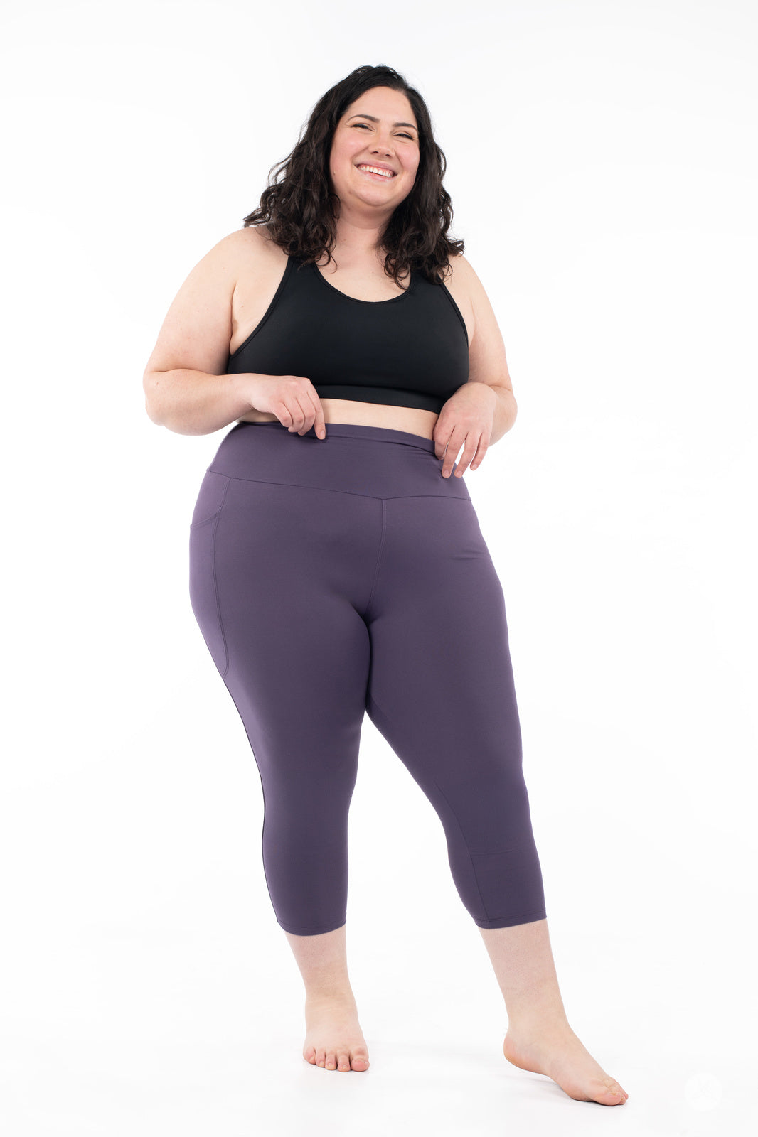 Smiling plus-size model showcasing plum SweetLegs High-Waist Pocket Crops with hands resting near waistband on white background