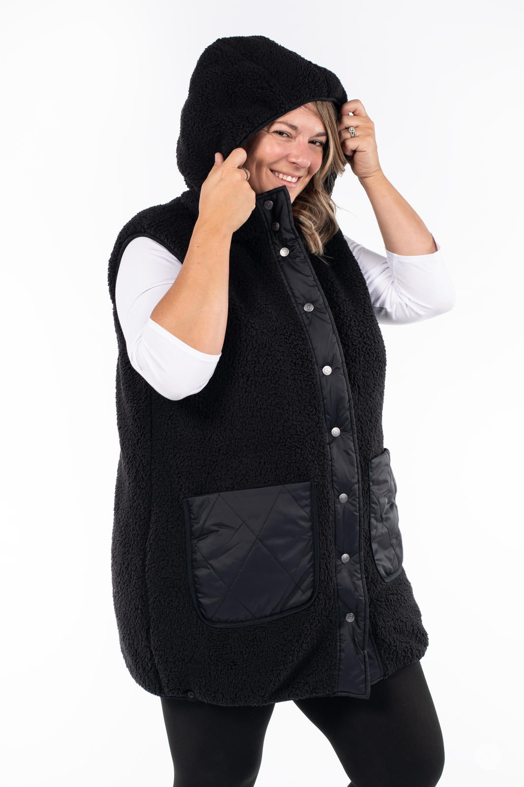 Smiling woman adjusting hood on black Reversible Quilted Sherpa Vest by SweetLegs Canada against white background