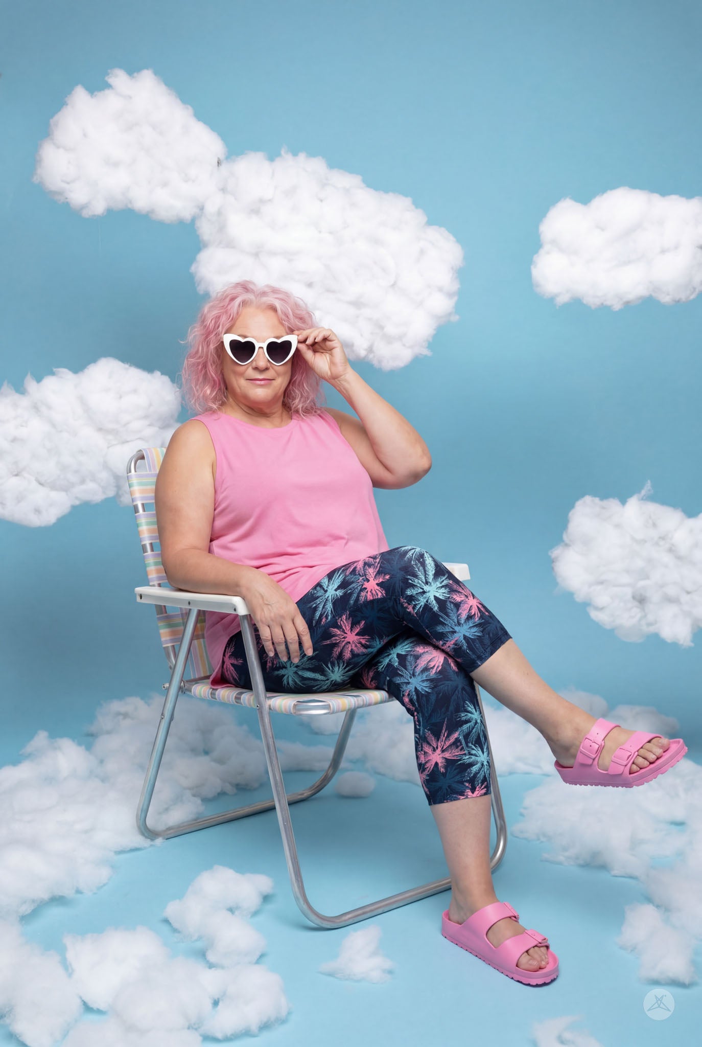 Woman with pink hair wearing SweetLegs Tiki Nights Crops with palm print, pink top, and heart-shaped sunglasses sitting in chair