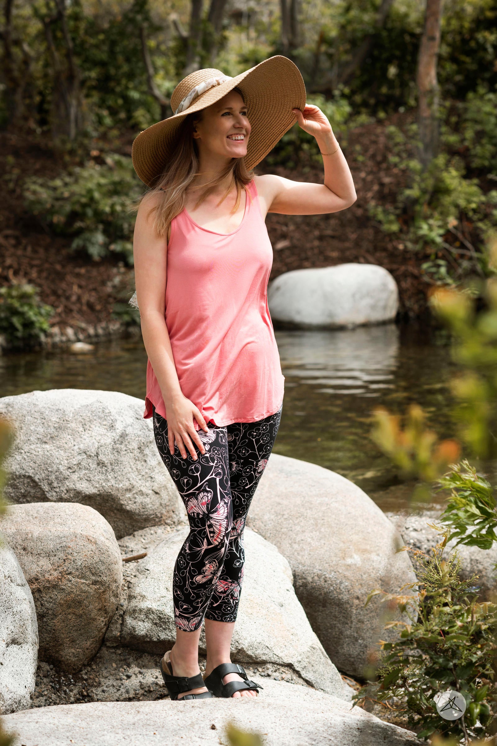 Woman wearing SweetLegs Butterfly Bliss 2.0 Crops with pink butterfly print paired with a coral tank top and large sun hat outdoors