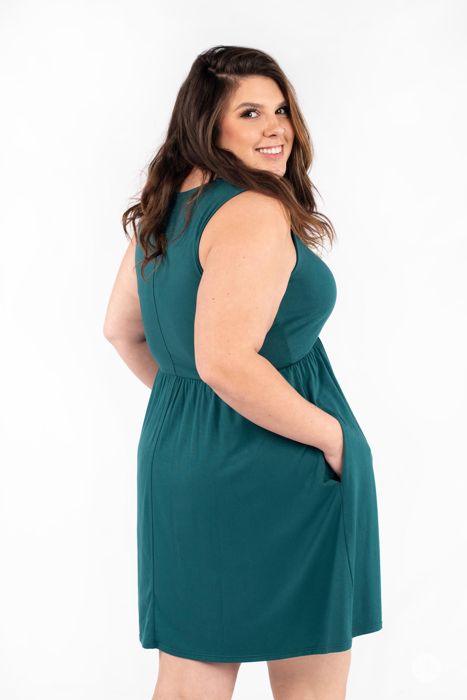 Plus-size model smiling over shoulder wearing SweetTops The Weekender Dress in teal with pockets on white background