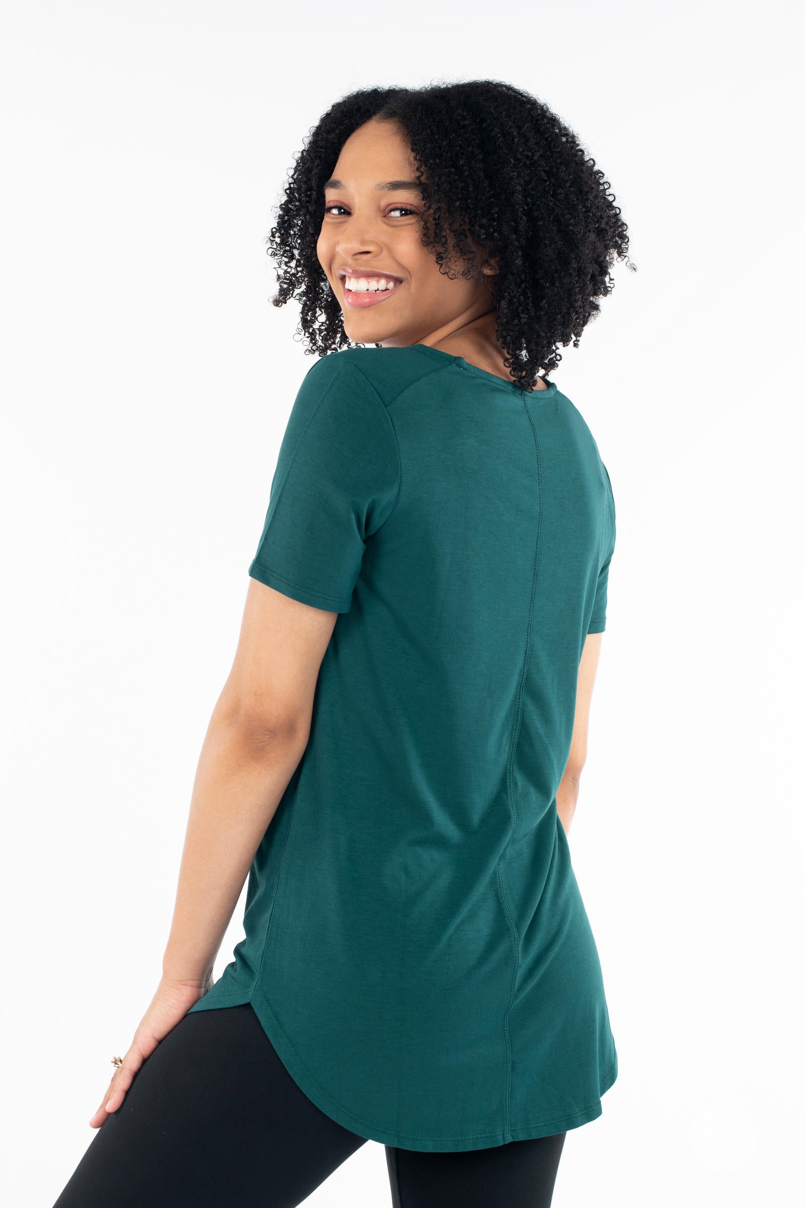 Smiling model wearing dark green Classic Tee by SweetLegs Canada with short sleeves and curved high low hem back view