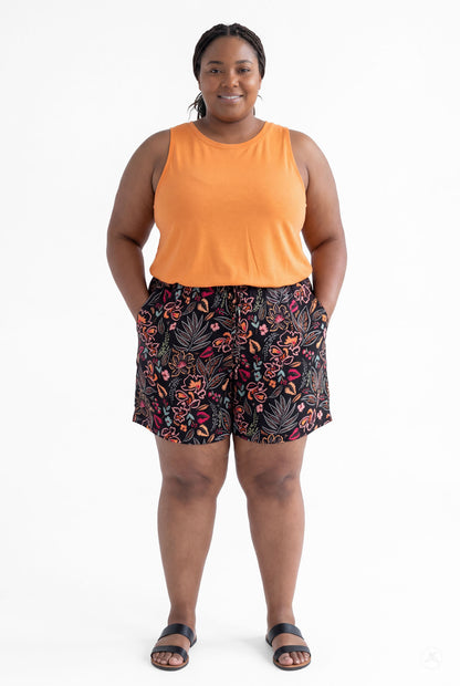 Plus size model wearing SweetLegs Breeze Shorts with floral print and orange sleeveless top, hands in pockets, smiling