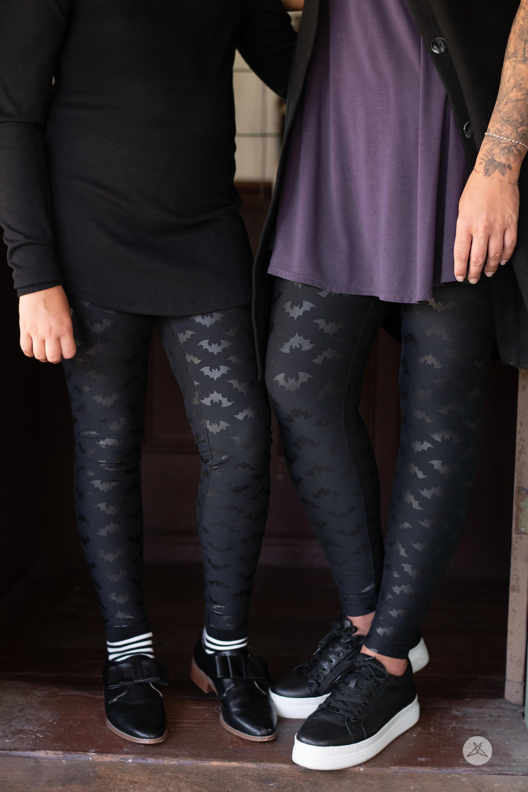 Two people wearing SweetLegs Flight Risk Kids leggings with glossy bat silhouettes on black fabric in doorway setting
