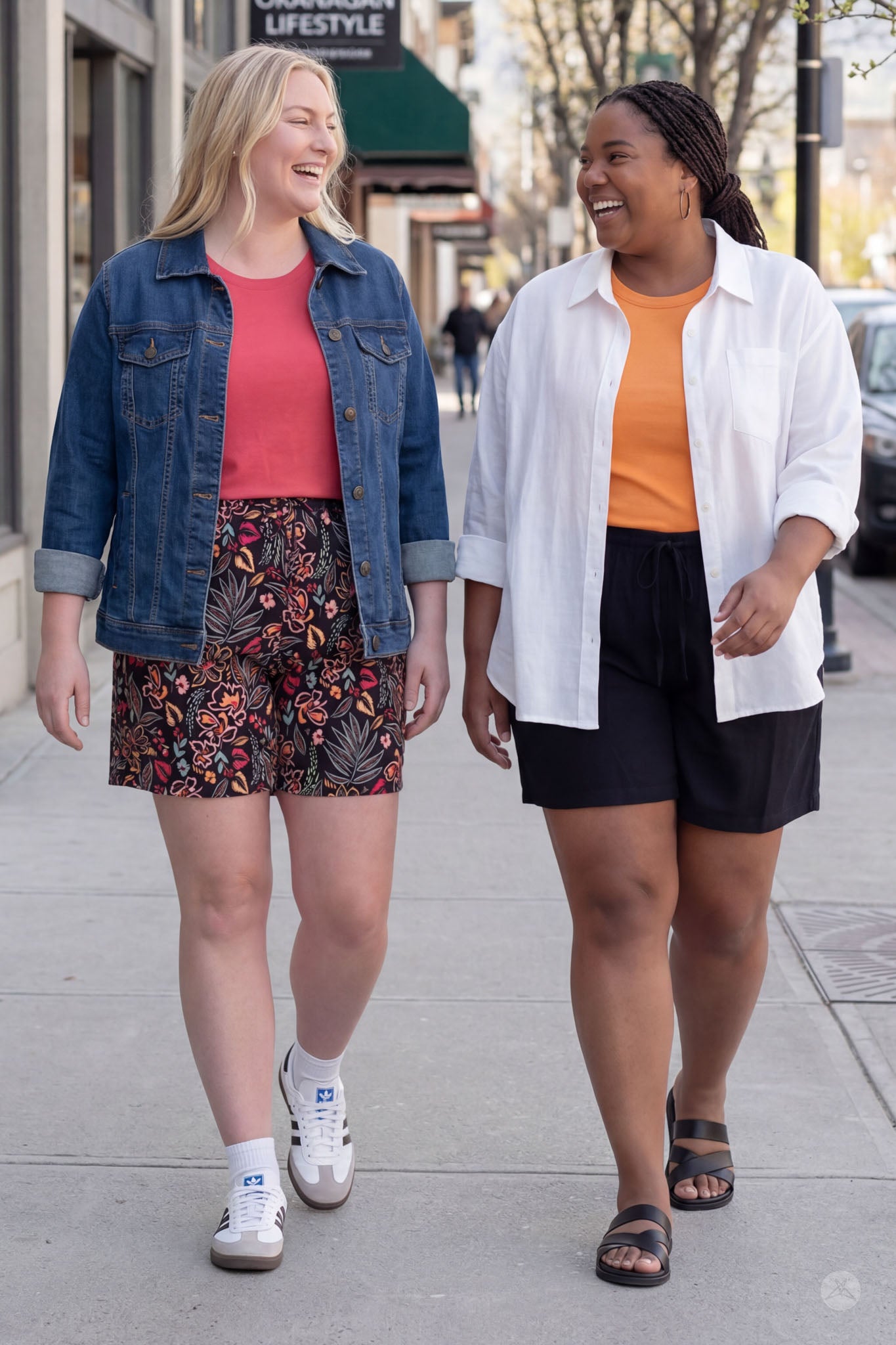 Two women walking outdoors wearing SweetLegs Breeze Shorts, one in black with orange top and white shirt, the other floral shorts with denim jacket