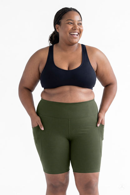 Smiling plus size model wearing SweetLegs Cedar High-Waisted Pocket Biker Shorts in green with black sports bra front view