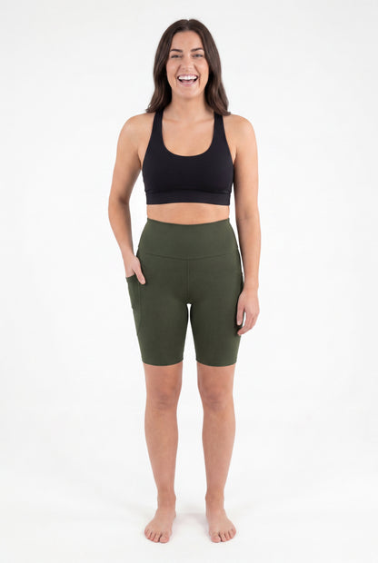 Smiling woman wearing SweetLegs Cedar High-Waisted Pocket Biker Shorts in green with black sports bra front view barefoot