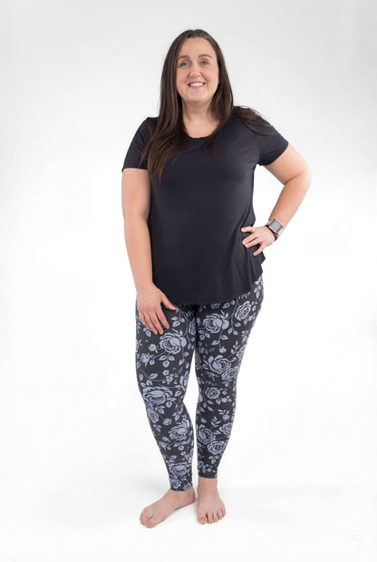 Woman wearing SweetLegs Shadow Garden leggings with soft grey-blue roses on a dark background and a black top