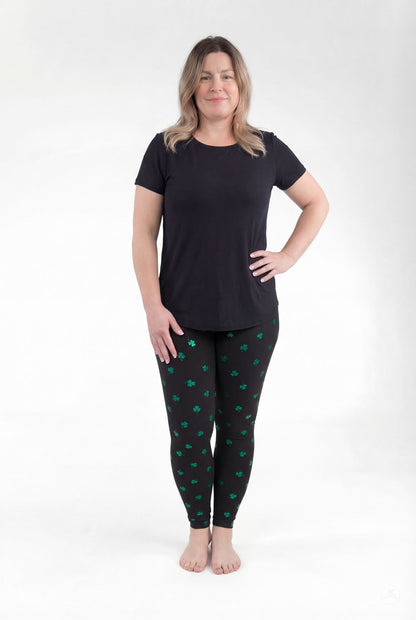 Woman modeling SweetLegs Lucky Glow leggings with green shamrock print on black background standing barefoot