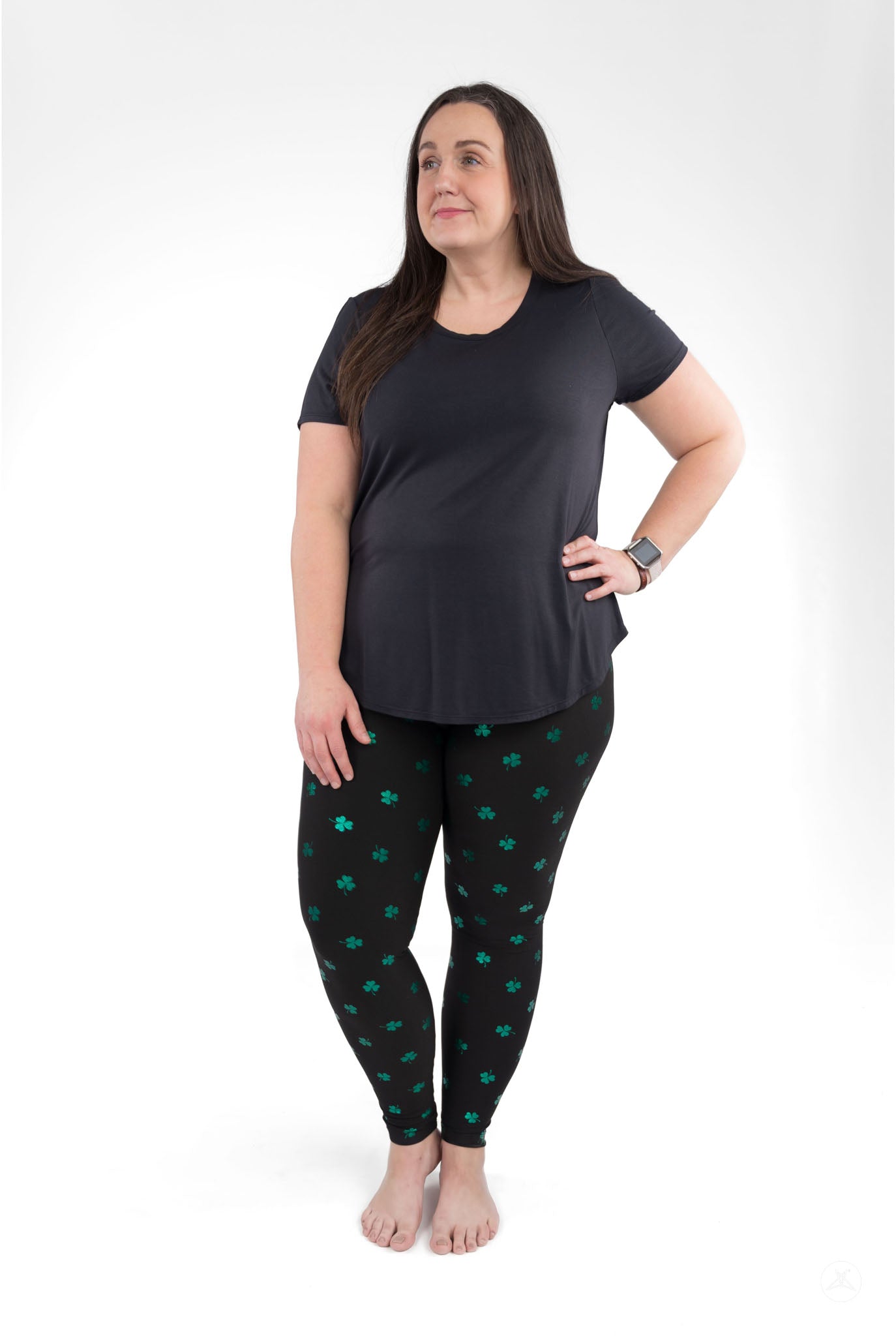 Woman wearing Lucky Glow leggings by SweetLegs with emerald shamrock print on black background, standing barefoot and smiling