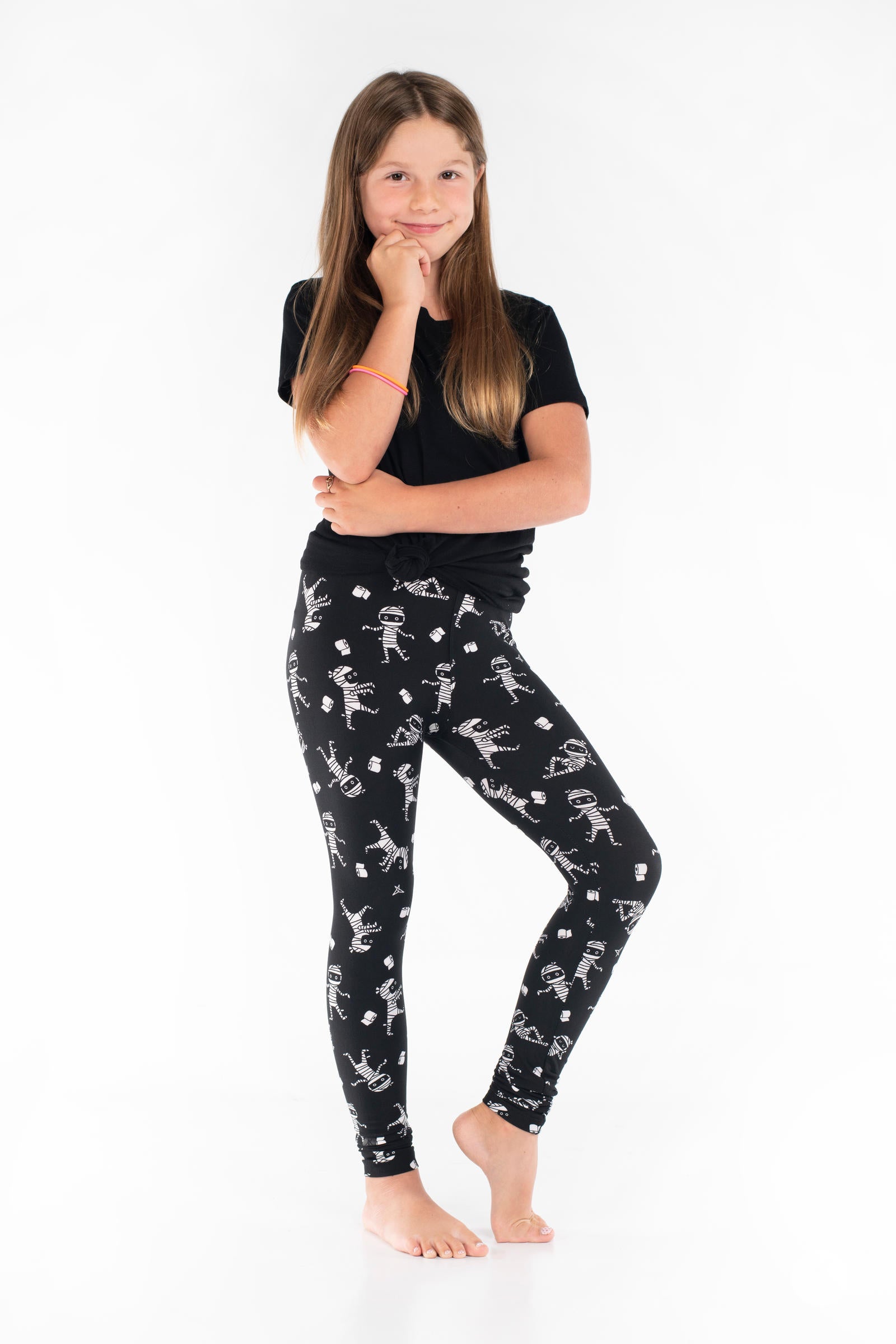 Under Wraps Kids leggings by SweetLegs with black fabric and white dancing mummy designs on smiling girl posing barefoot
