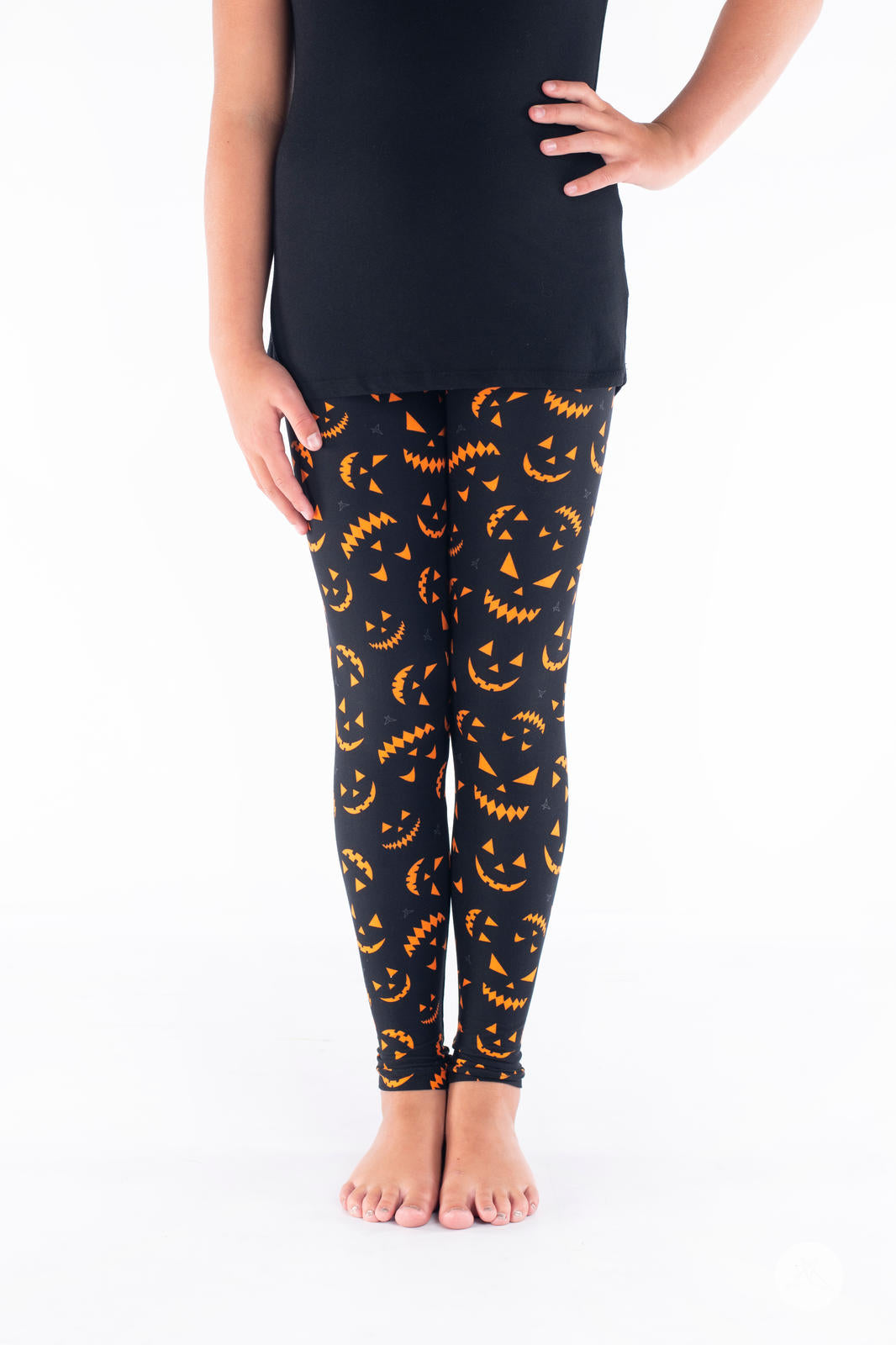 Child wearing SweetLegs Creep It Real Kids leggings with bright orange jack-o-lantern faces on black background standing barefoot